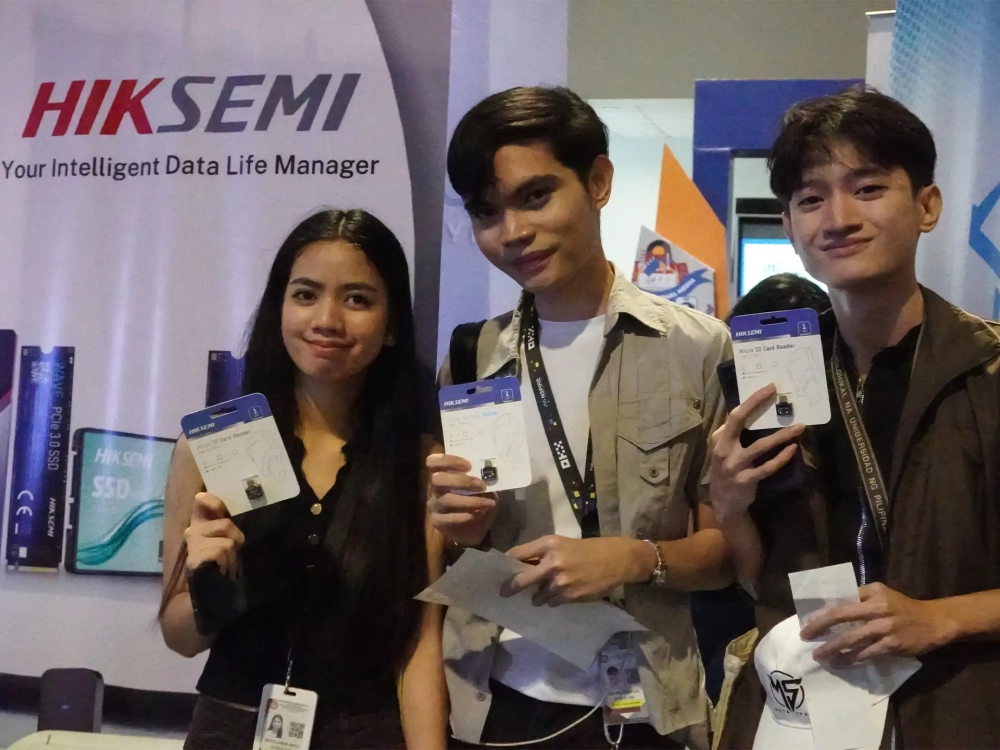 Three event attendees smiling while holding up packaged Hiksemi Micro SD Card Readers in front of a brand banner.